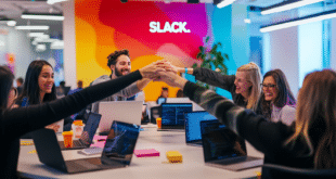 Plusieurs personnes se joignent les mains au milieu d'une table, équipé d'ordinateurs portables, et un grand panneau au mur indique « SLACK ».