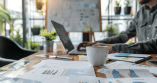 Un entrepreneur est devant son ordinateur, dans son bureau, avec plusieurs papiers financiers sur la table et une verre