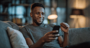 Homme dans la trentaine exprimant une joie discrète après avoir gagné un pari sportif, assis dans son salon avec un téléphone à la main.